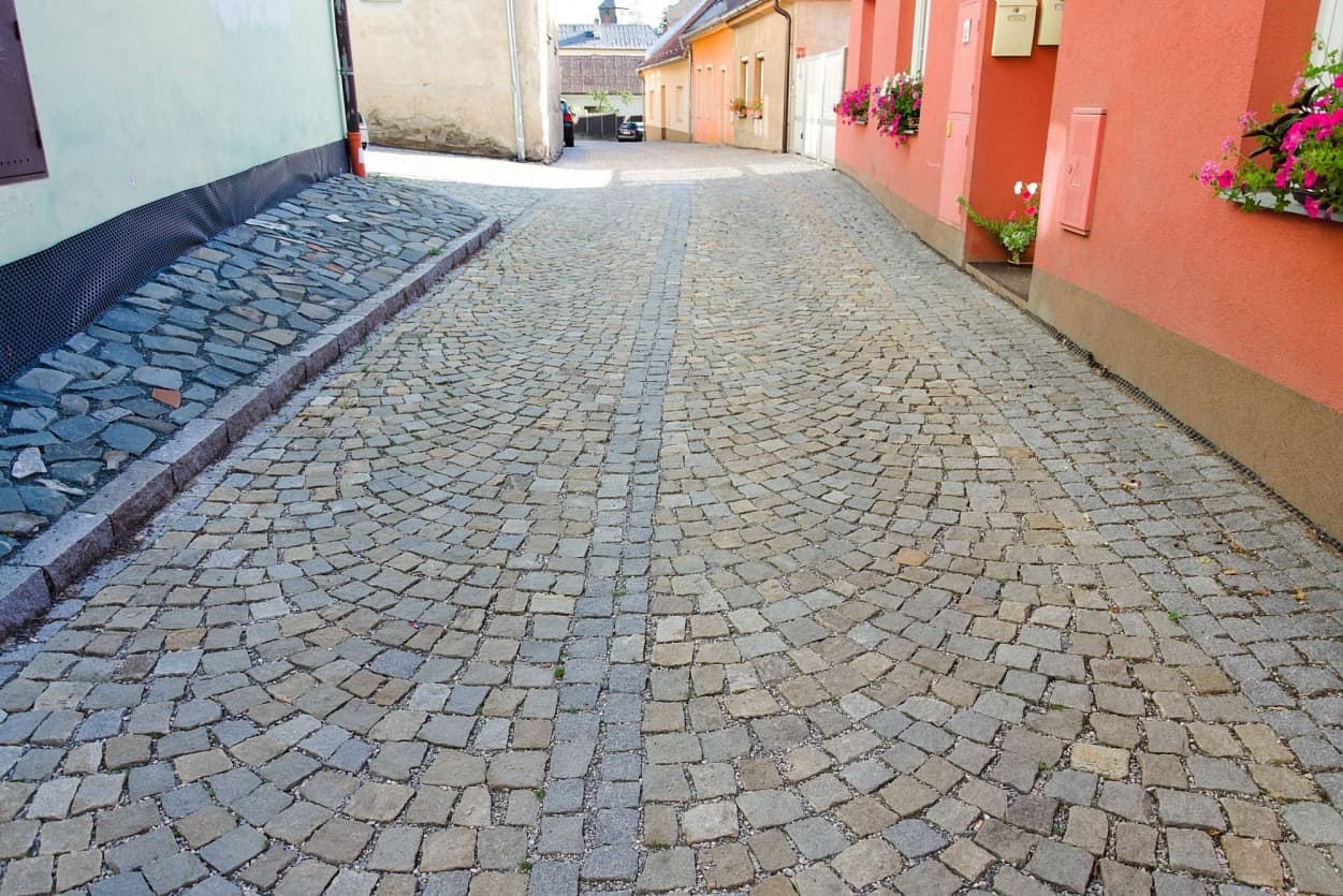 Dlážděná ulička s šedožlutými žulovými kostkami, lemovaná domy s květinami v oknech a zvýšeným chodníkem z lomového kamene.