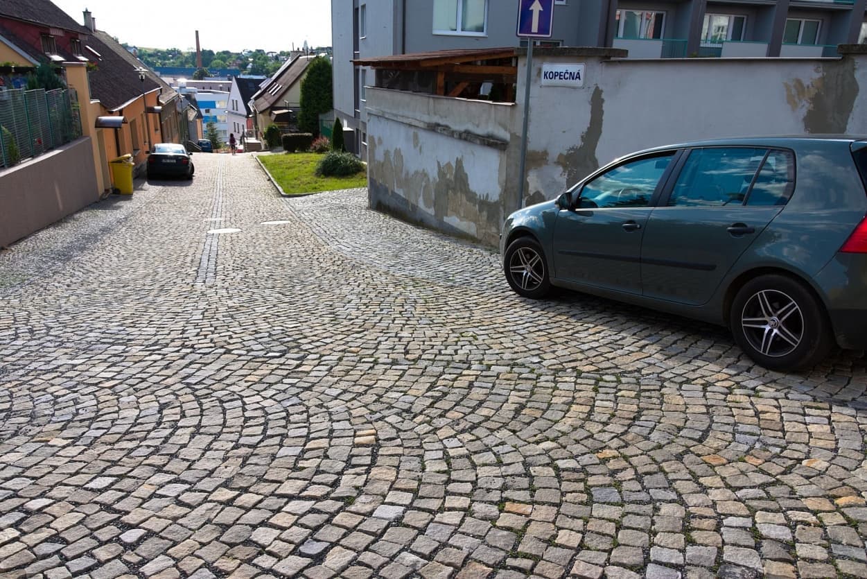 Dlážděná ulice se šedožlutými štípanými kostkami, prudkým sklonem a zaparkovaným autem, lemovaná domy a zdí s cedulí „Kopečná“.