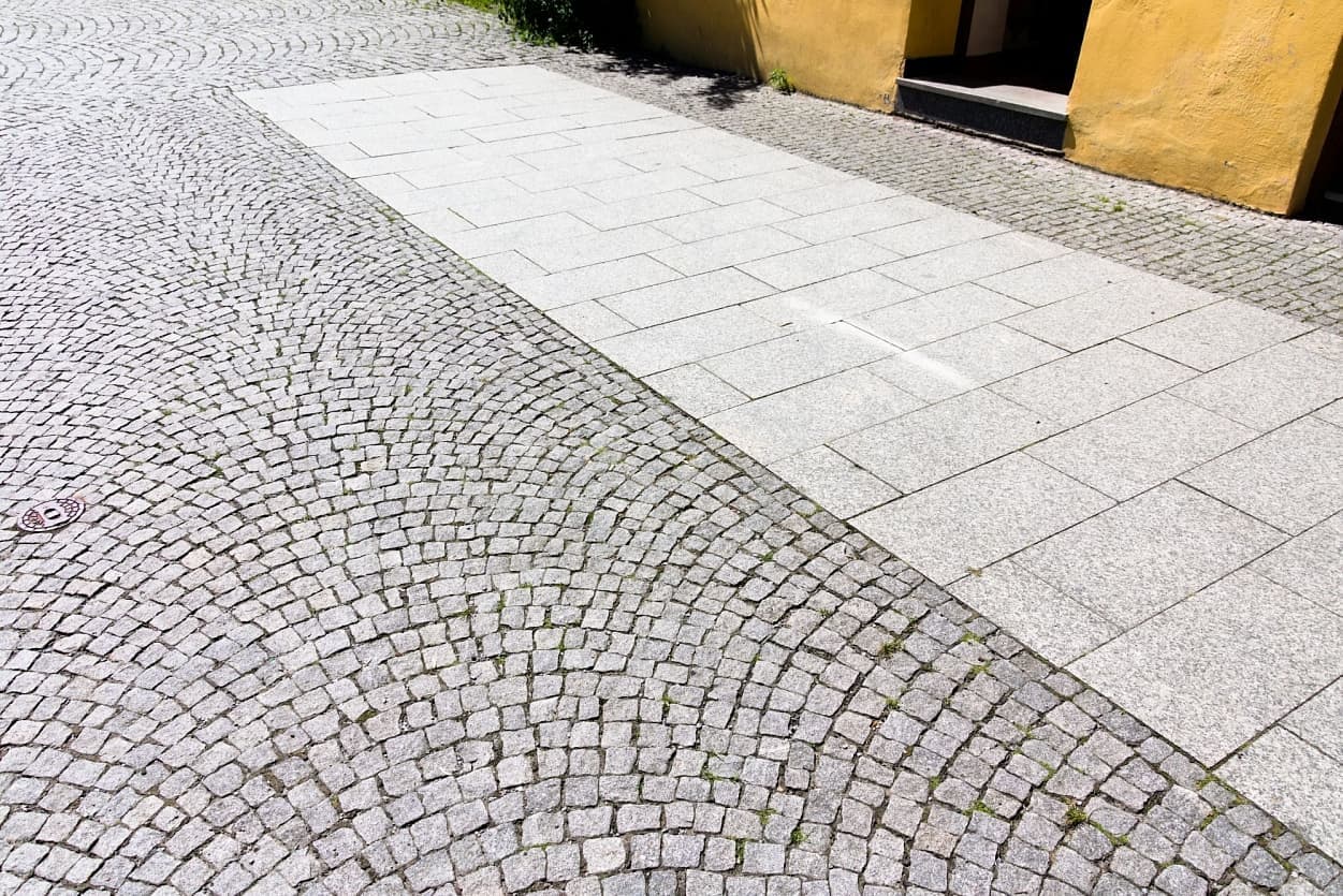 Šedá žulová dlažba s kombinací štípaných kostek a formátovaných desek, vytvářející kontrastní vzor v městském prostředí.