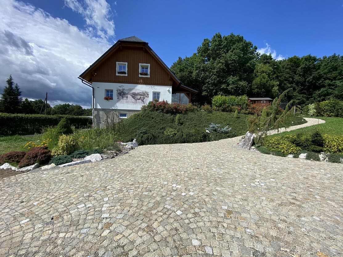 Přírodní kamenná dlažba ze žulových kostek ve žlutém odstínu, tvořící příjezdovou cestu k domu s dřevěným obkladem.