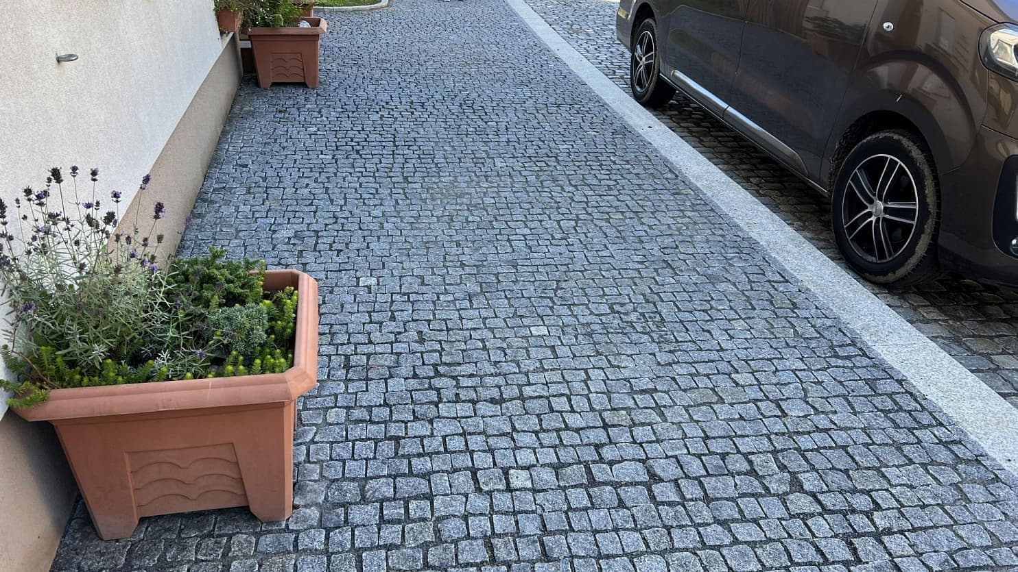 Chodník a parkovací plocha ze šedých žulových kostek, ohraničené řezaným obrubníkem, doplněné květináči s rostlinami.