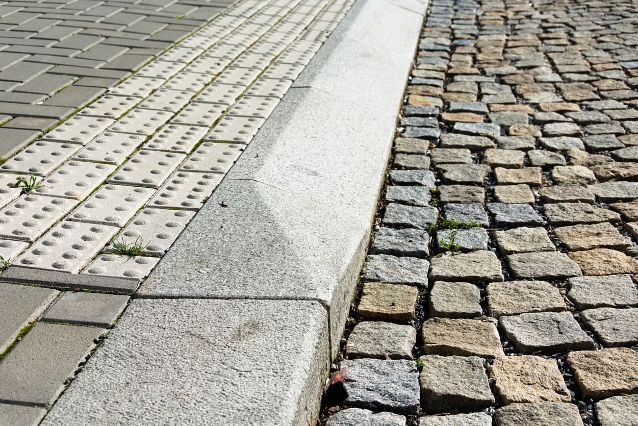 Detailní pohled na přechod mezi chodníkem s hmatovou dlažbou a silnicí z žulových kostek, oddělených masivním žulovým obrubníkem.