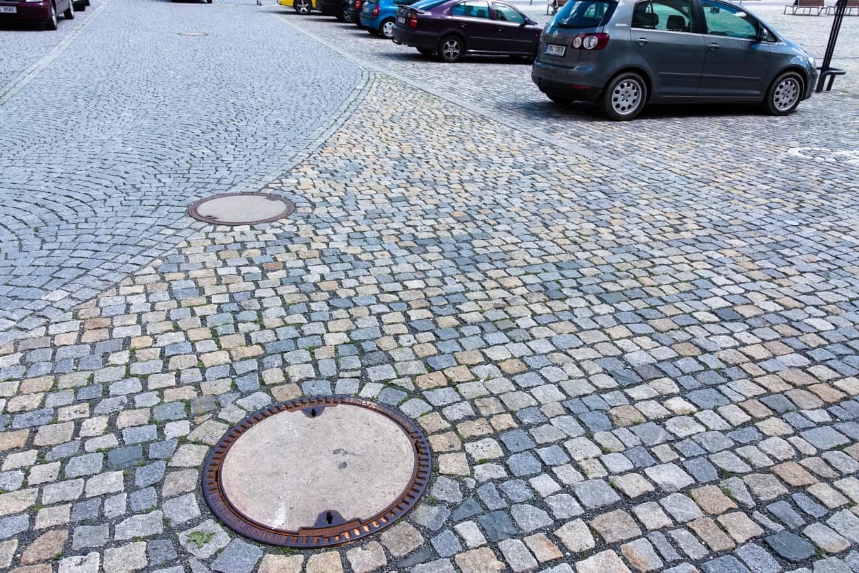 Dlážděná plocha z žulových kostek na náměstí s kanalizačními poklopy a parkovanými auty v pozadí.