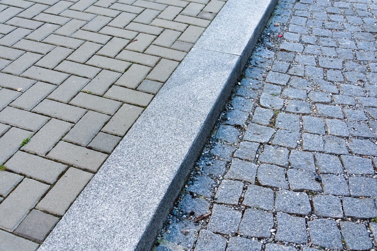 Pohled na přechod mezi chodníkem z betonové dlažby a silnicí s šedými žulovými kostkami, oddělený masivním obrubníkem.