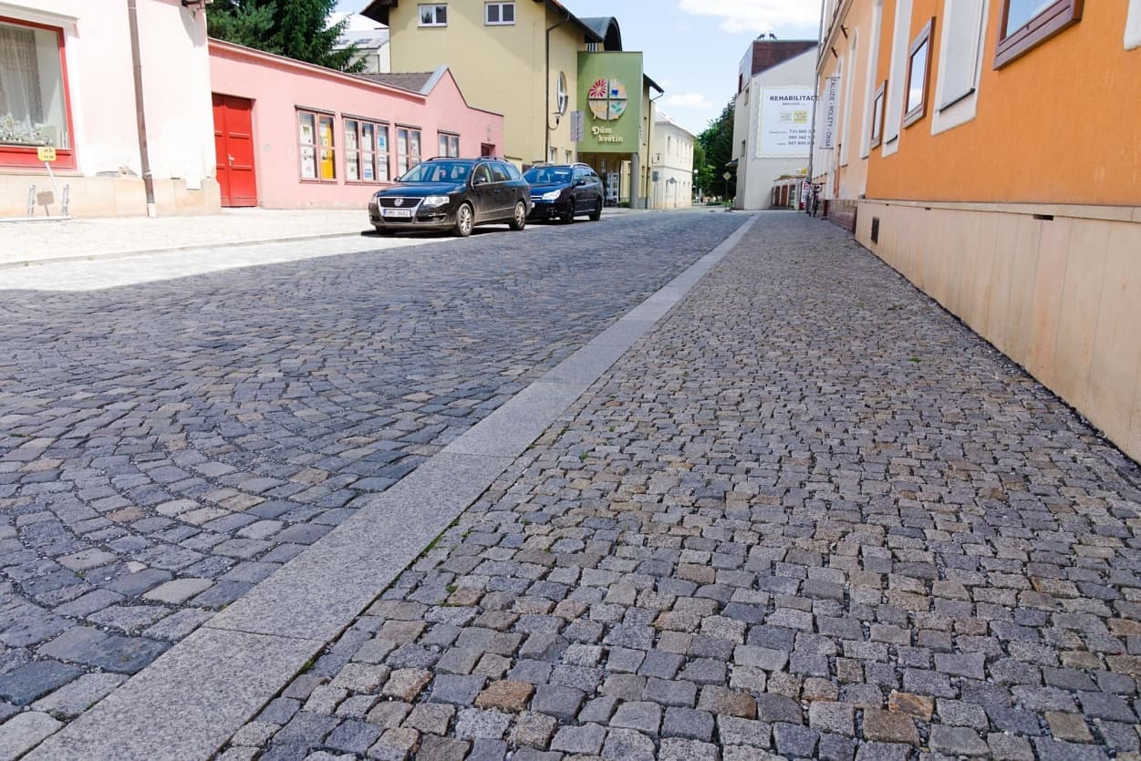 Ulice s dlažbou z žulových kostek, doplněná žulovým obrubníkem a parkovanými auty podél chodníku.