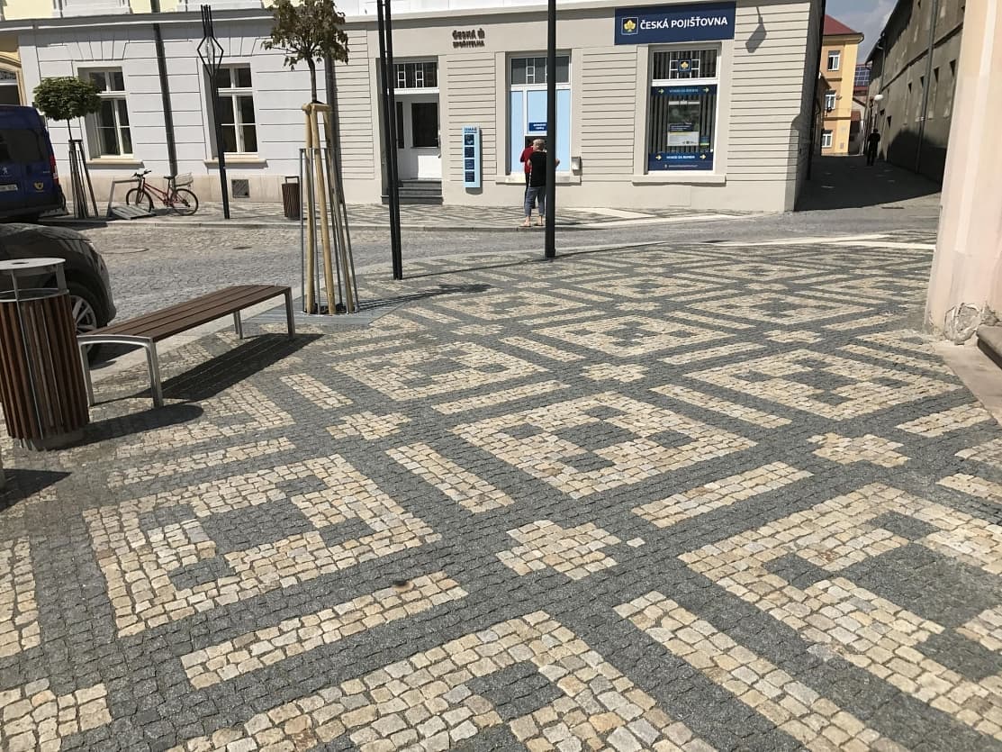 Ornamentální dlažba z žulových kostek na náměstí, doplněná lavičkami, odpadkovými koši a mladými stromy.