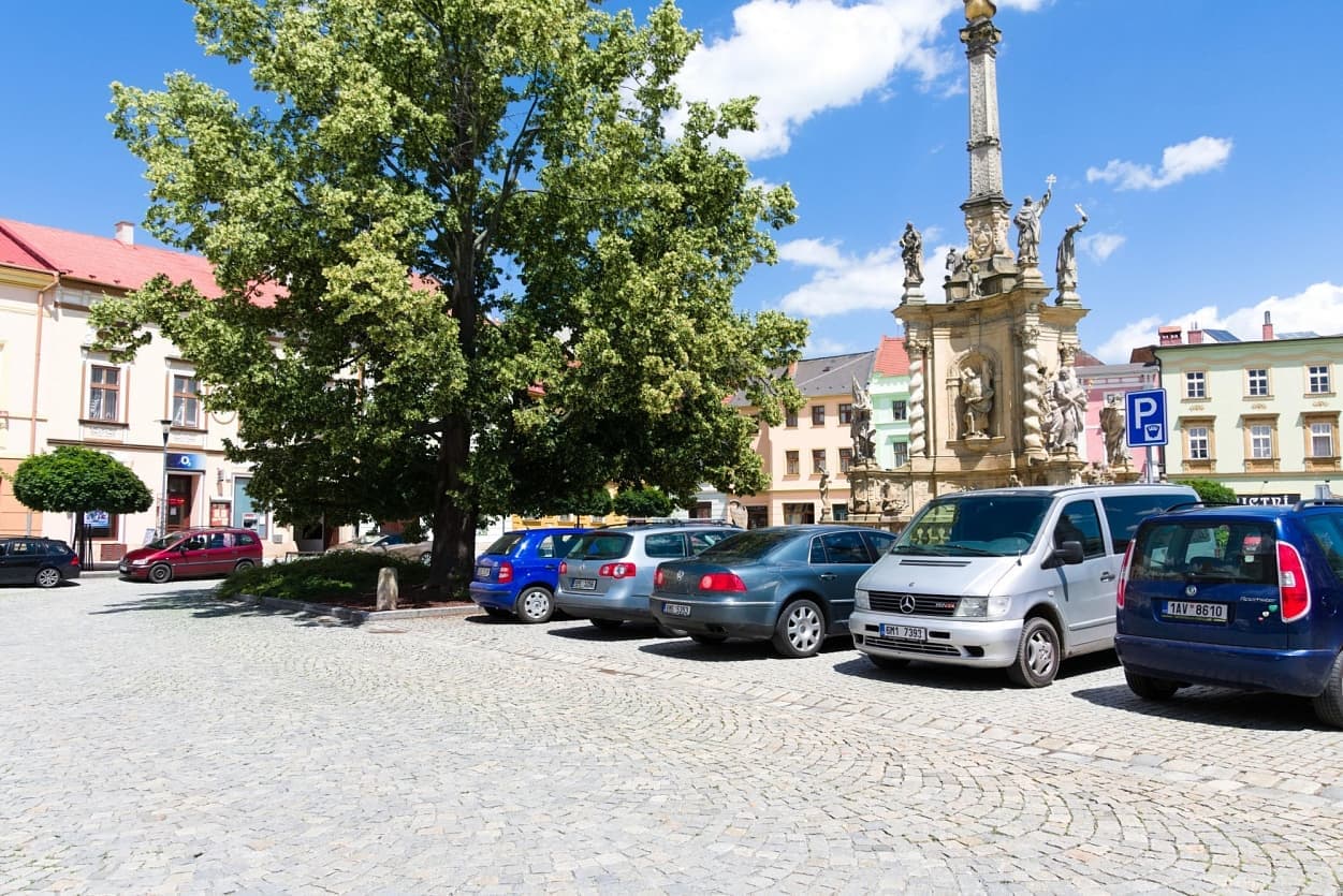 Historické náměstí v Uničově s žulovou dlažbou, barokním sloupem a parkovanými automobily pod vzrostlým stromem.