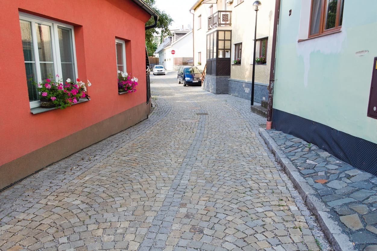 Úzká dlážděná ulička se žulovou dlažbou, lemovaná barevnými domy s květinami v oknech a zvýšeným chodníkem z lomového kamene.