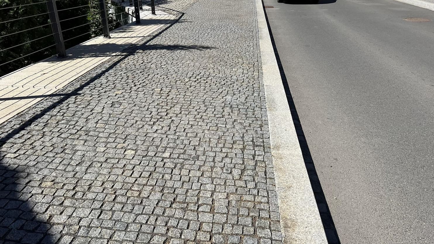 Chodník ze žulových kostek v šedém odstínu, lemovaný obrubníky, harmonicky ladí s moderním zábradlím a asfaltovou vozovkou.