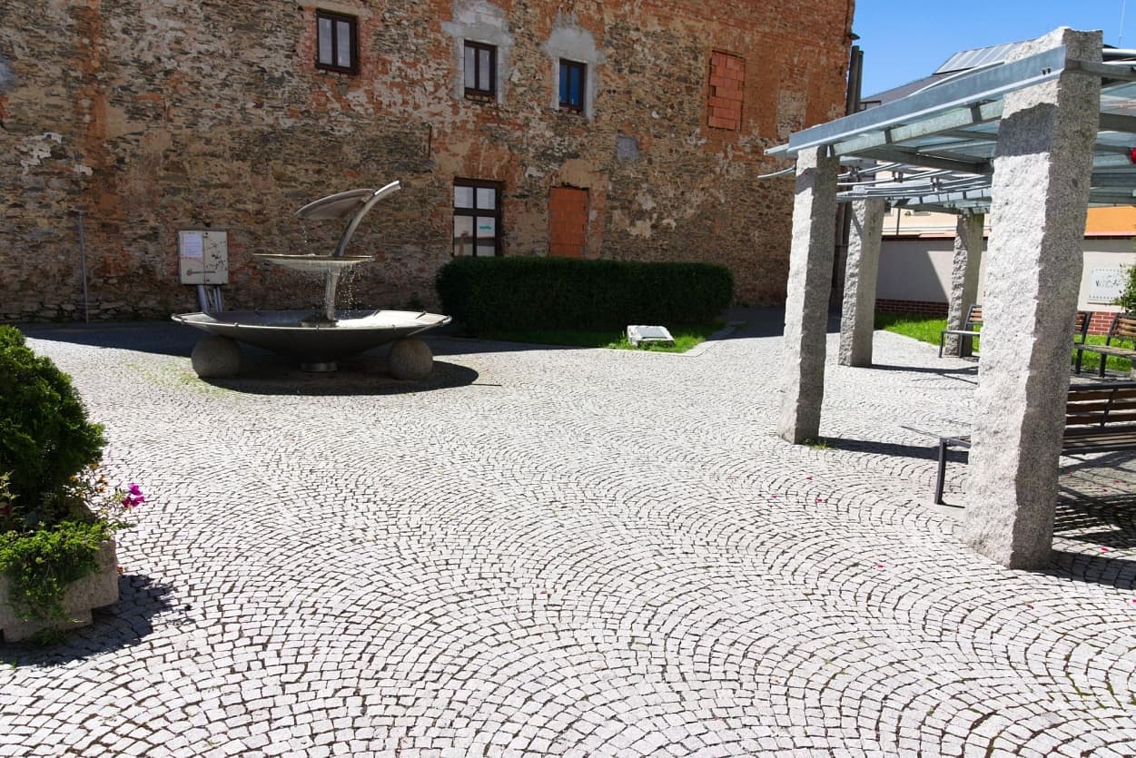 Vějířová žulová dlažba s moderní fontánou a žulovými sloupky v historickém městském prostoru.