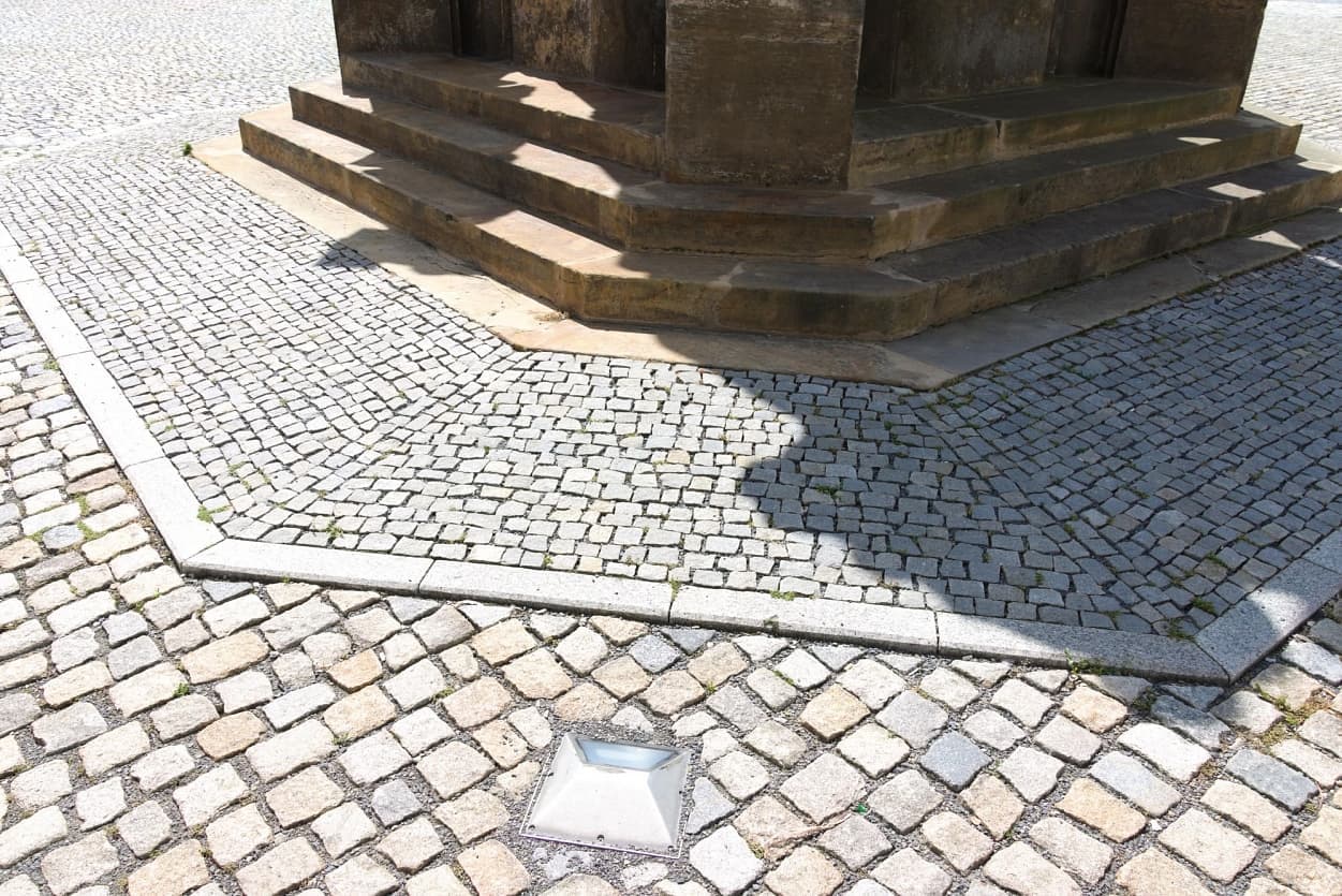Detail historického kamenného podstavce kašny na náměstí s žulovou dlažbou a obrubníky v různých formátech.