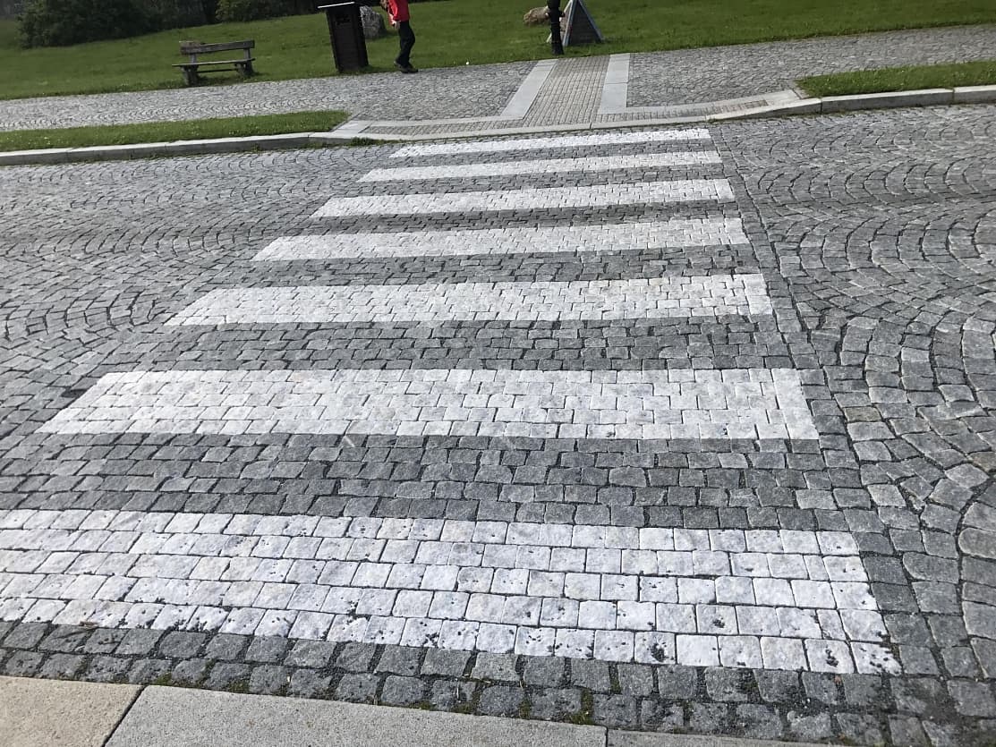 Přechod pro chodce z bílého mramoru a tmavé žulové kostky v Karlově Studánce, obklopený přírodním kamenem.