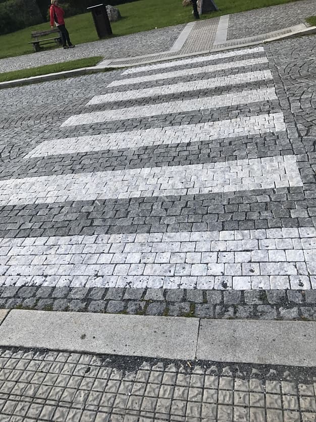 Přechod pro chodce z bílého mramoru a tmavé žulové dlažby v Karlově Studánce, obklopený přírodním kamenem.