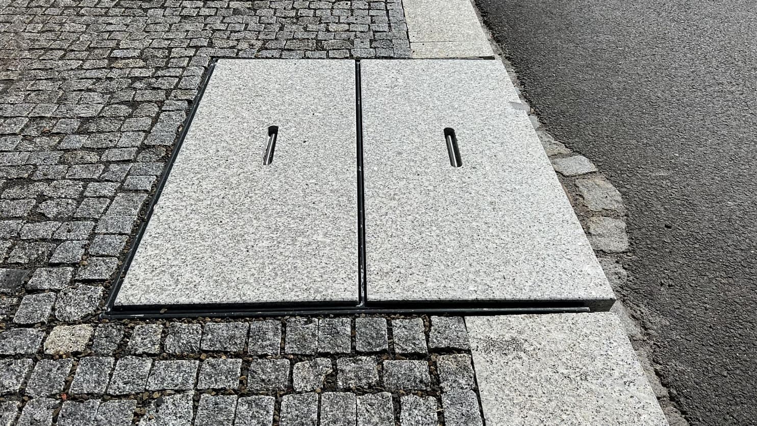 Žulový poklop z šedé žuly, zasazený do chodníku z žulových kostek, zajišťuje odolné zakrytí technické infrastruktury.