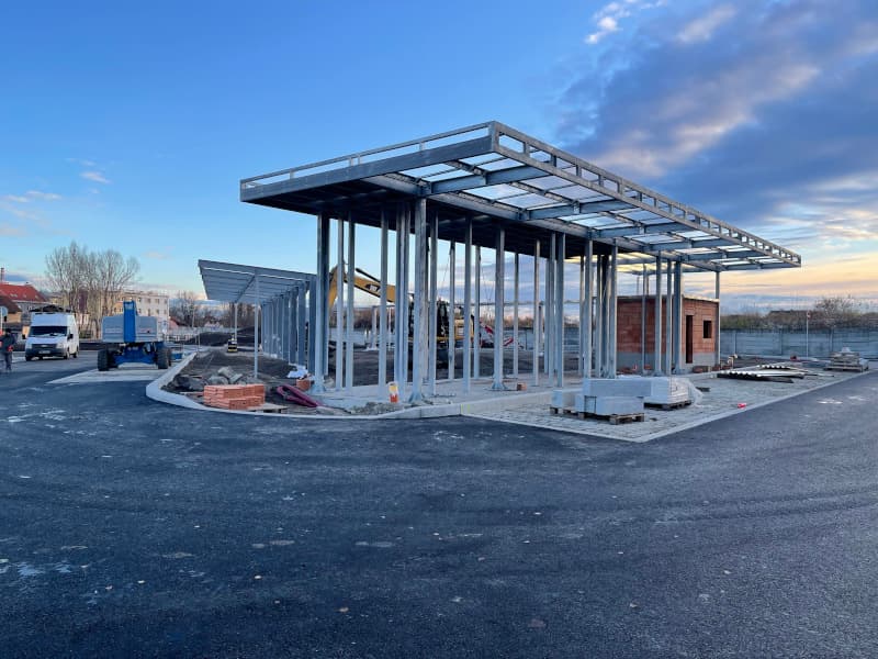 Stavba autobusového terminálu v Kralupech nad Vltavou, ocelová konstrukce přístřešku, asfaltový povrch a stavební materiál na místě.