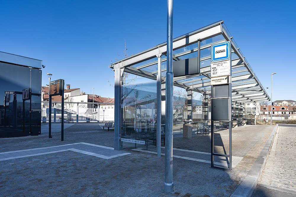 Moderní autobusový terminál v Kralupech nad Vltavou s proskleným přístřeškem, informační tabulí, žulovou dlažbou a lavičkami.