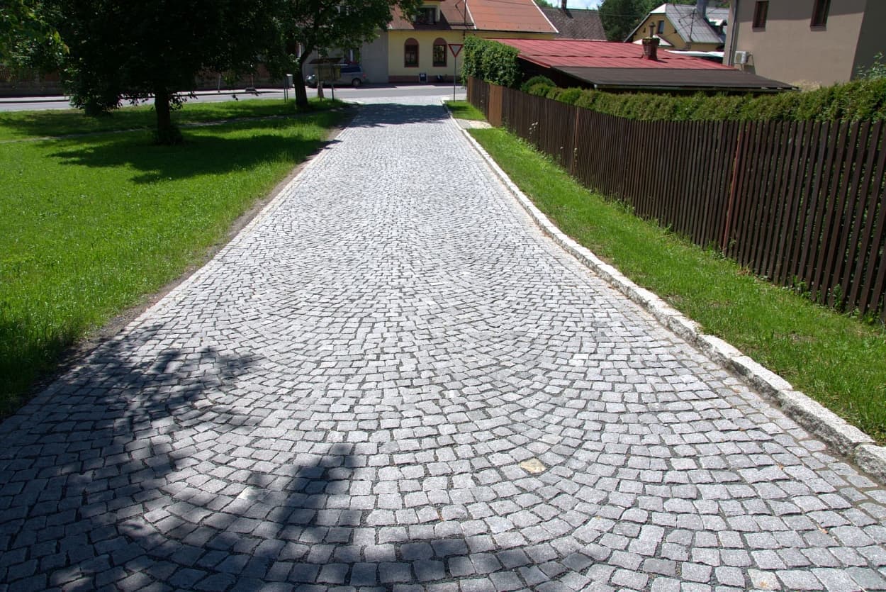Cesta vydlážděná šedou štípanou žulovou kostkou v obloukovém vzoru, lemovaná žulovými obrubníky a obklopená zelení.
