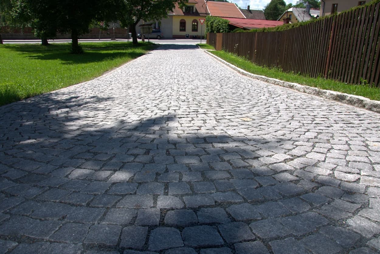 Cesta ze šedé štípané žulové kostky s obloukovým vzorem, doplněná žulovými obrubníky, lemovaná zelení a dřevěným plotem.