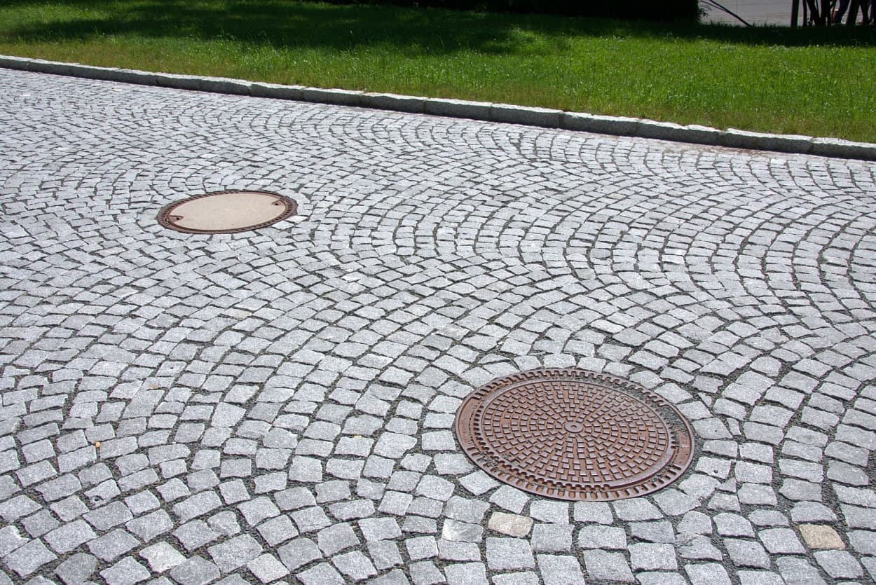 Žulová štípaná dlažba v obloukovém vzoru s kanalizačními poklopy, lemovaná žulovými obrubníky a obklopená zeleným trávníkem.