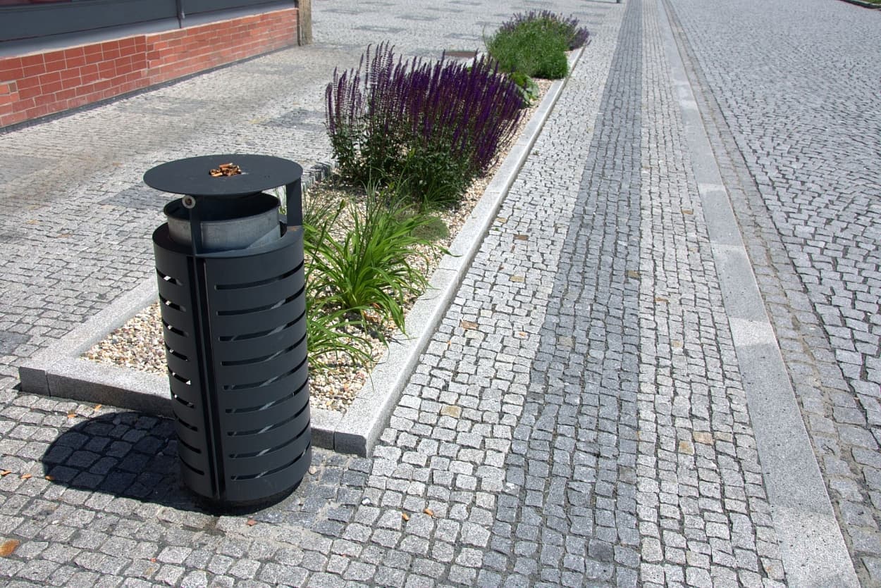Žulová štípaná dlažba s odvodňovacím pásem, žulovými obrubníky a zeleným ostrůvkem s květinami, doplněná odpadkovým košem.