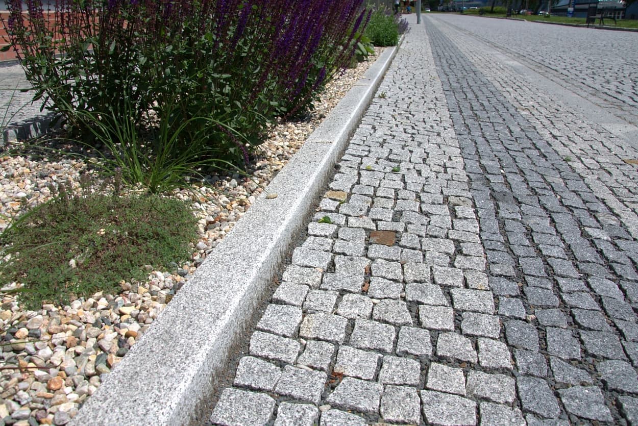Detail žulového obrubníku oddělujícího štípanou žulovou dlažbu od květinového záhonu se štěrkem a kvetoucími rostlinami.