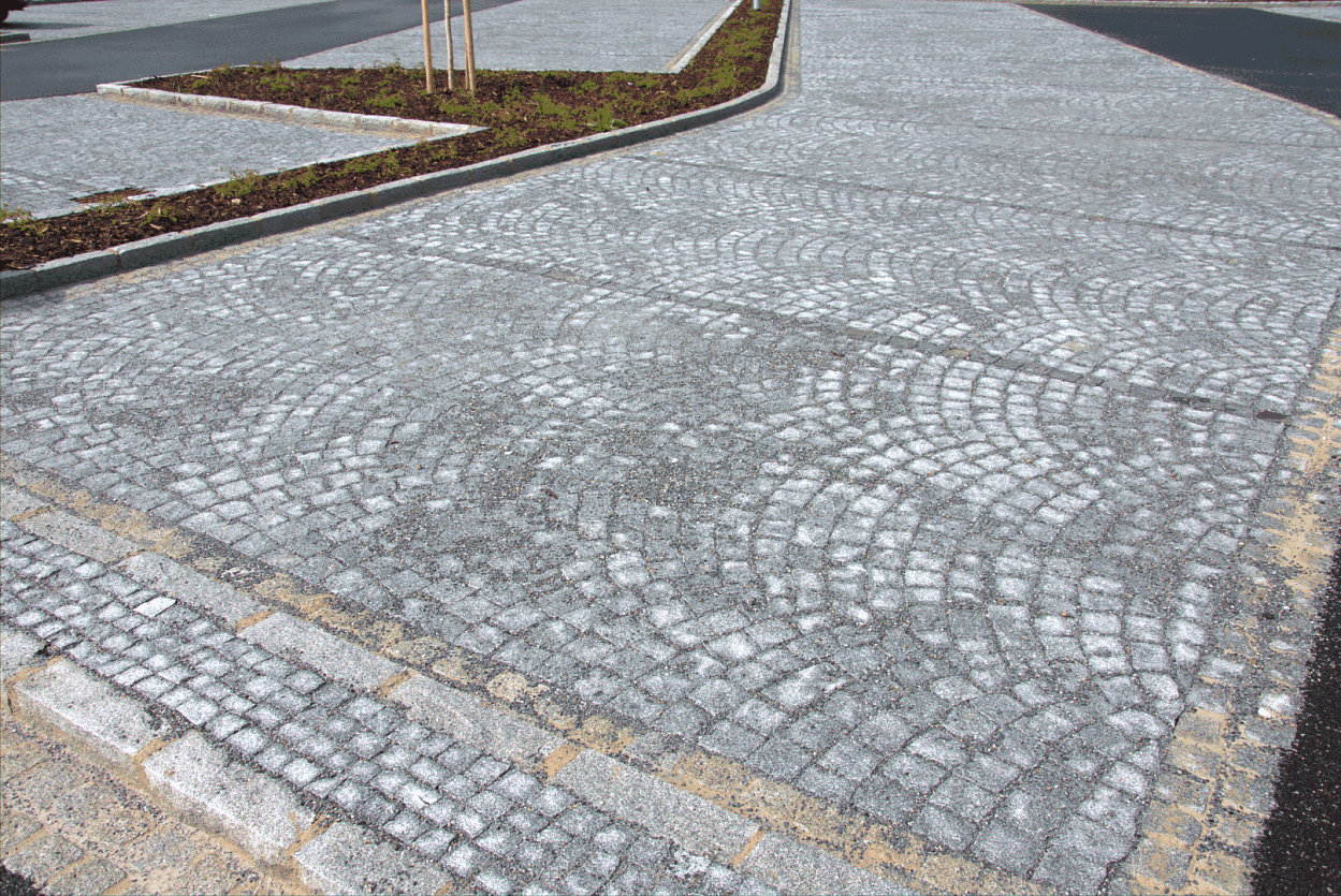 Parkovací plocha vydlážděná žulovou štípanou kostkou ve vějířovém vzoru, lemovaná žulovými obrubníky a doplněná ostrůvky.