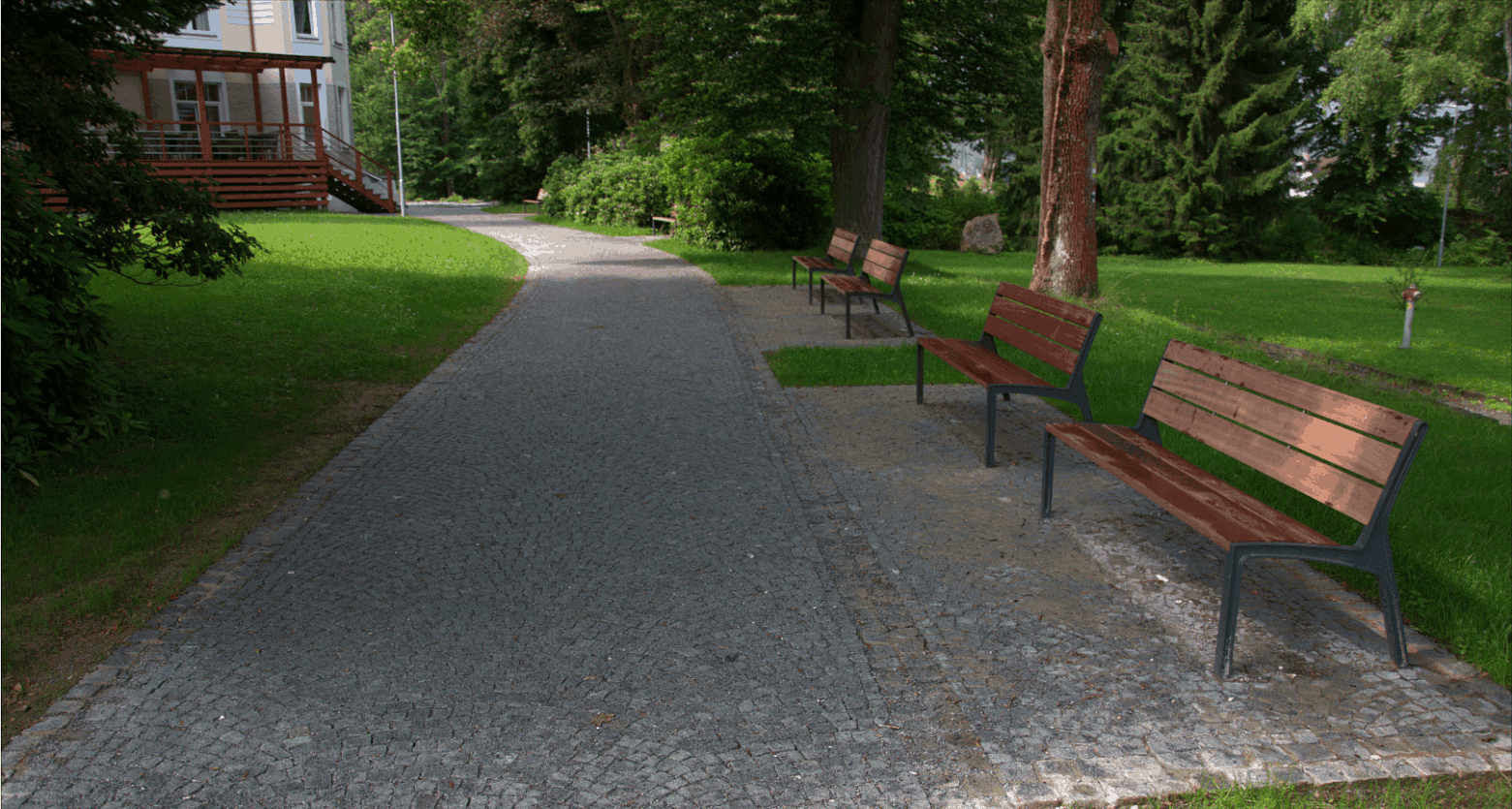 Parková cesta vydlážděná žulovou štípanou kostkou, lemovaná lavičkami a zelení, vytvářející příjemné místo k odpočinku.