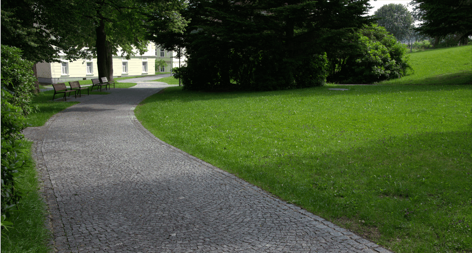 Meandrující parková cesta ze žulové štípané kostky, lemovaná zelení, vytvářející klidné místo k procházkám a relaxaci.