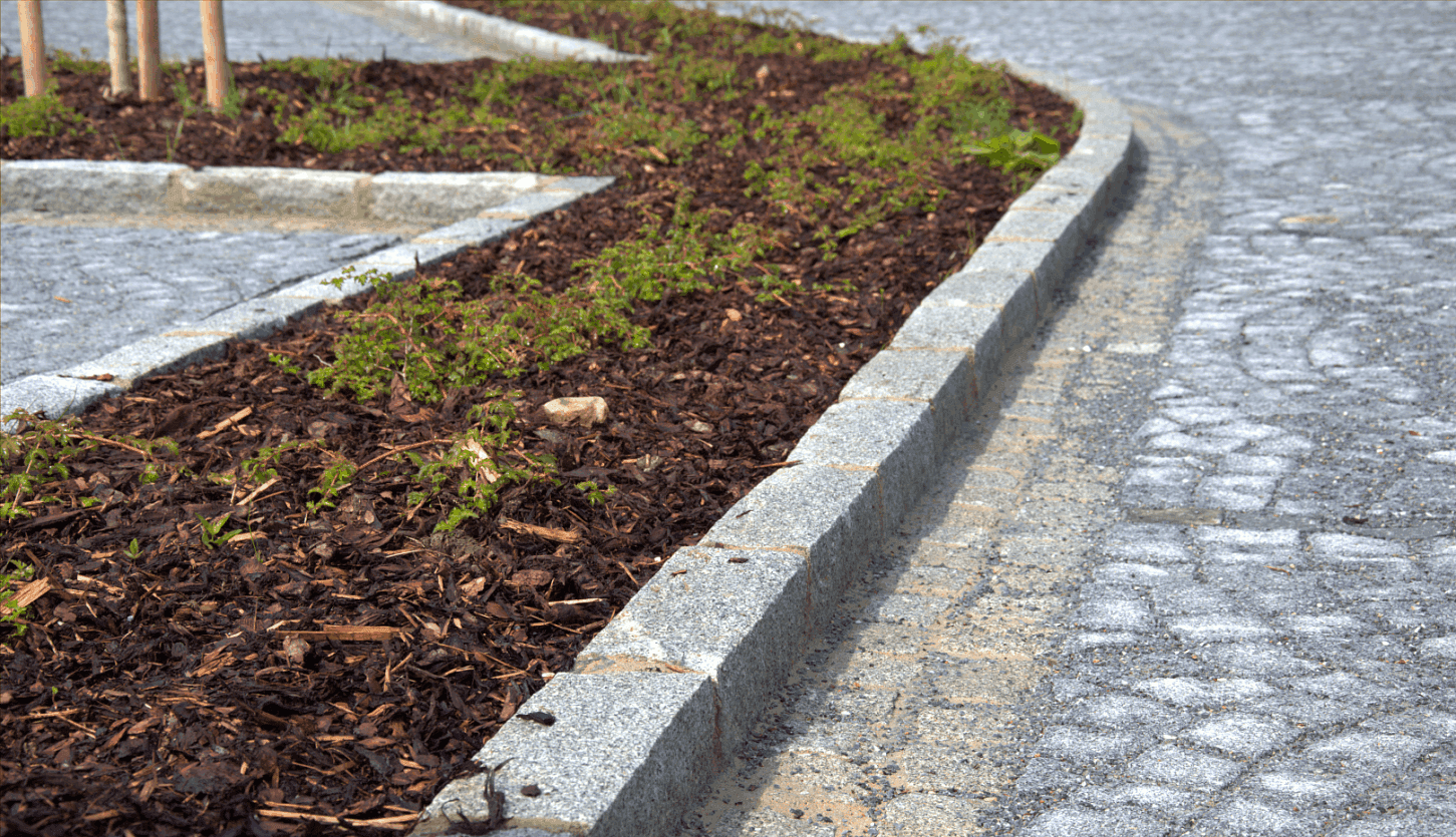 Detail žulového obrubníku a štípané dlažby u zeleného ostrůvku s čerstvě vysazenými rostlinami, odděleného mulčovací kůrou.