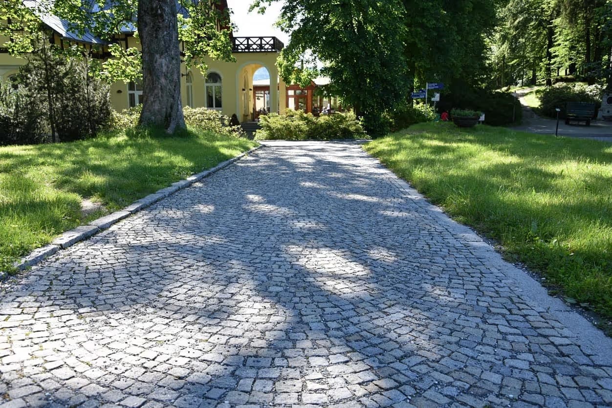 Chodník z obloukově kladené žulové dlažby v Priessnitzových léčebných lázních, lemovaný zelení a historickou budovou.