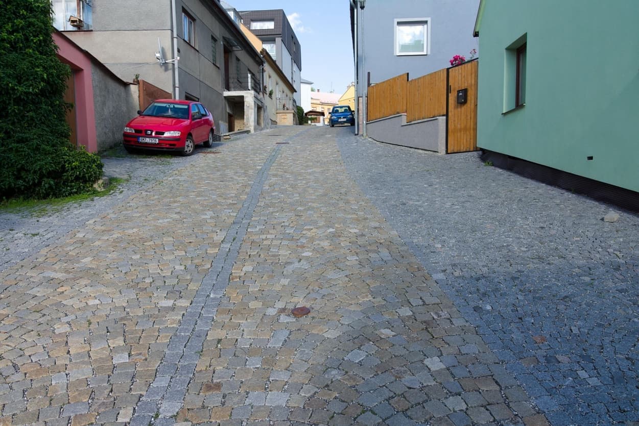 Úzká ulice dlážděná šedožlutými žulovými kostkami s centrálním odvodňovacím pásem, obklopená moderní i tradiční zástavbou.