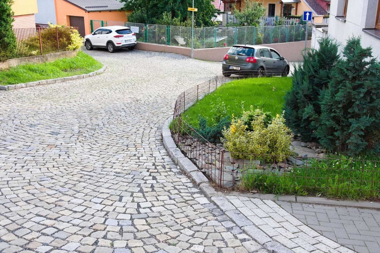 Zakřivená ulice dlážděná žulovými kostkami v šedožlutém odstínu s žulovým obrubníkem a zeleným předzahrádkovým prostorem.