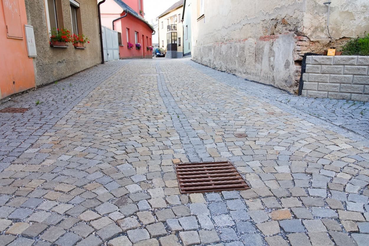 Historická ulice s dlažbou z žulových kostek v šedožlutém odstínu, doplněná středovým odvodňovacím pásem a litinovou mříží.