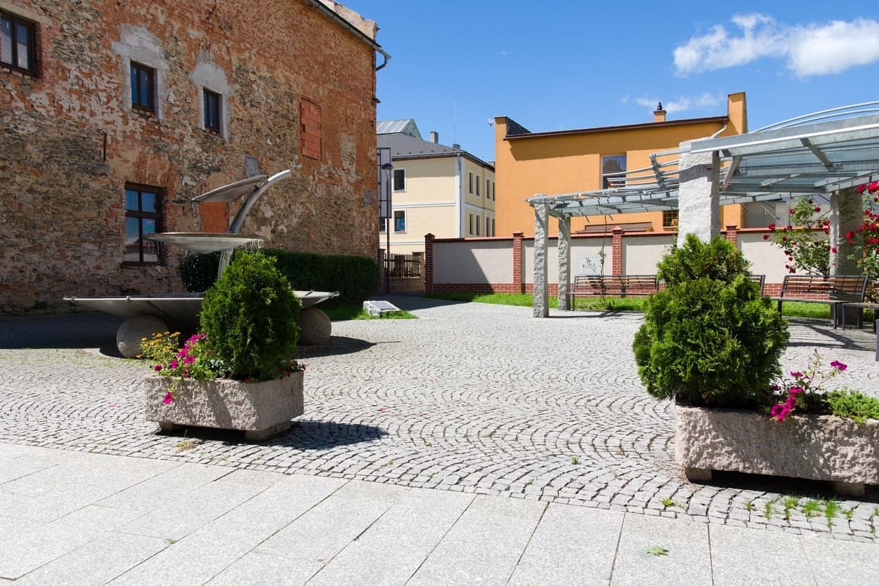 Vějířová žulová dlažba, moderní fontána a masivní žulové sloupky přístřešku v městském prostranství.
