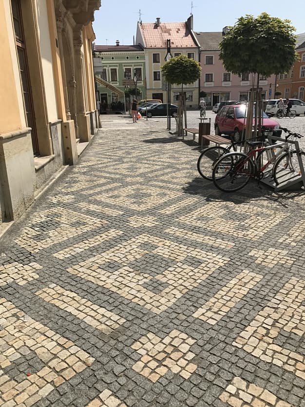 Chodník na náměstí s ornamentální dlažbou z přírodního kamene, lemovaný stromy, lavičkami a stojany na kola.