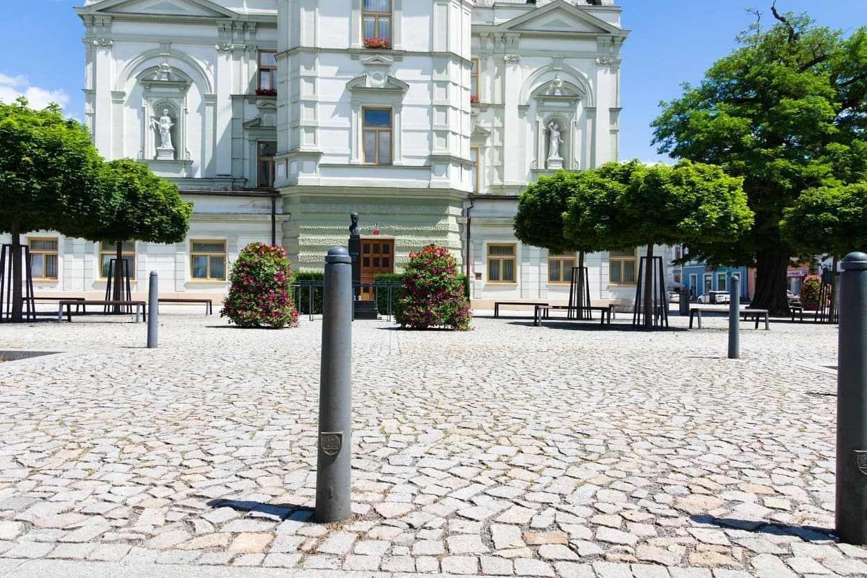 Náměstí před historickou budovou s vydlážděným povrchem z přírodního kamene a moderními sloupky.