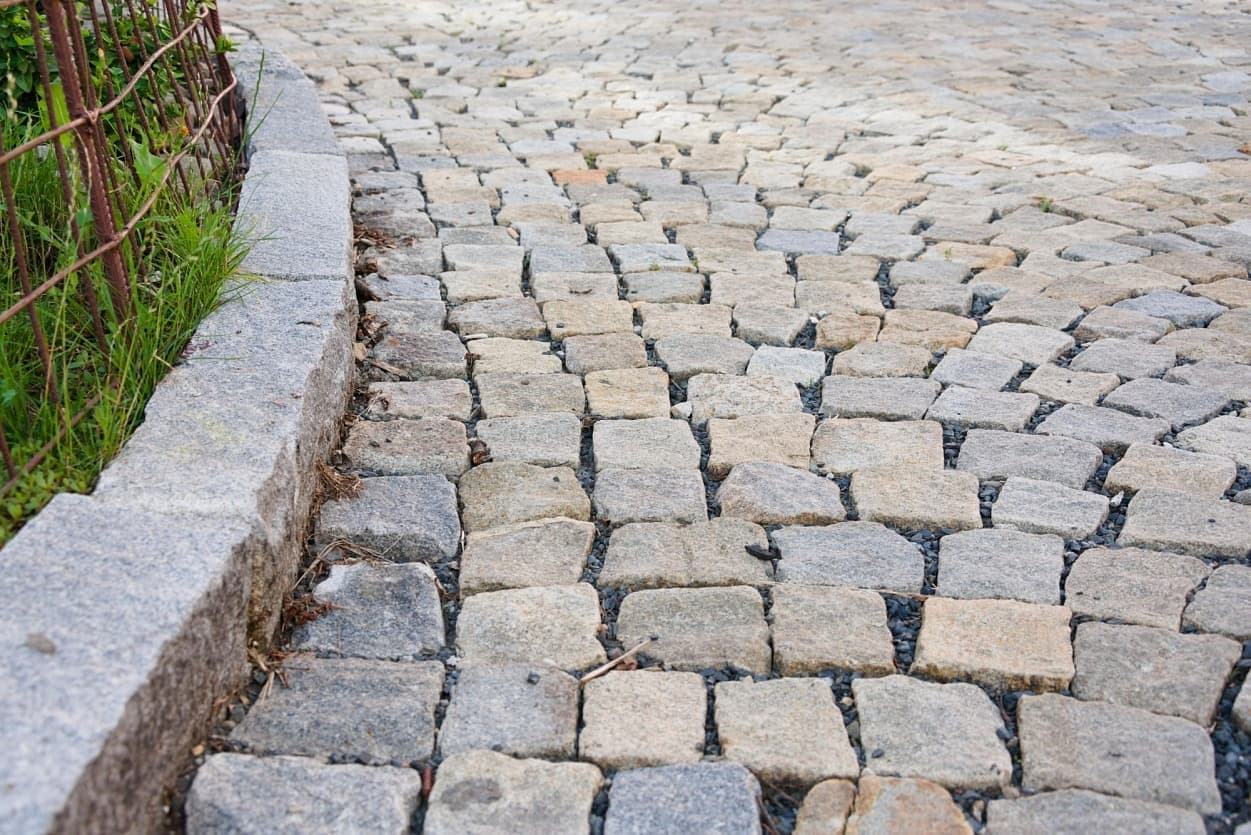 Detail šedožlutých žulových kostek s nepravidelným tvarem, lemovaných masivním žulovým krajníkem podél zeleného porostu