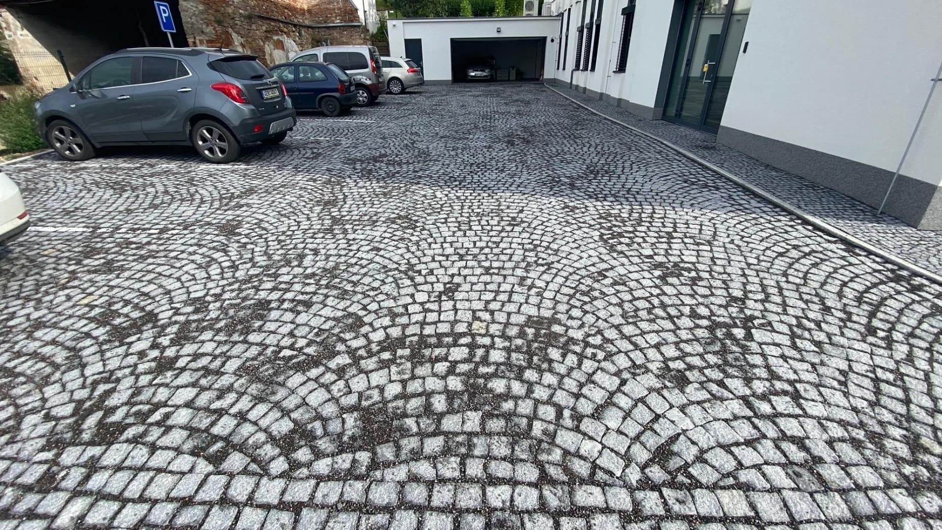 Parkoviště s obloukově kladenou dlažbou z tmavé žulové kostky, doplněné moderní budovou a garážovým vjezdem.
