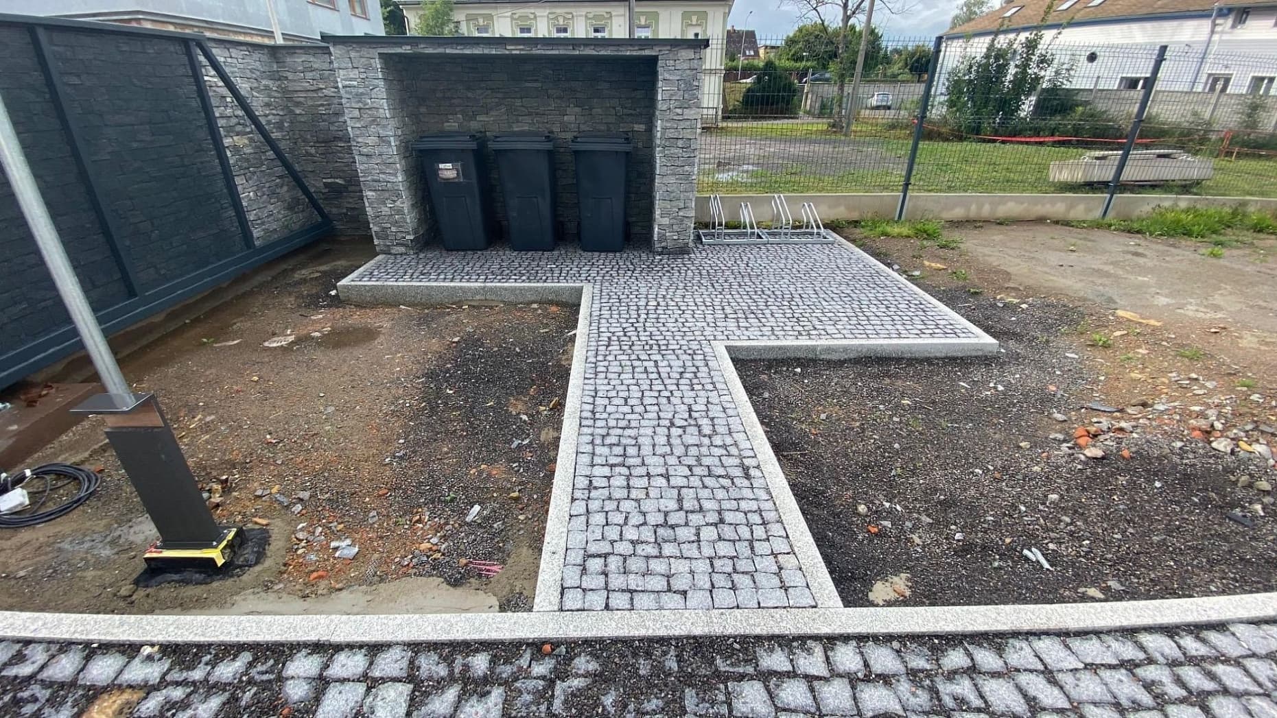 Chodníky a zpevněné plochy ze žulových kostek s obrubníky u přístřešku na popelnice, doplněné kamenným obkladem stěn.