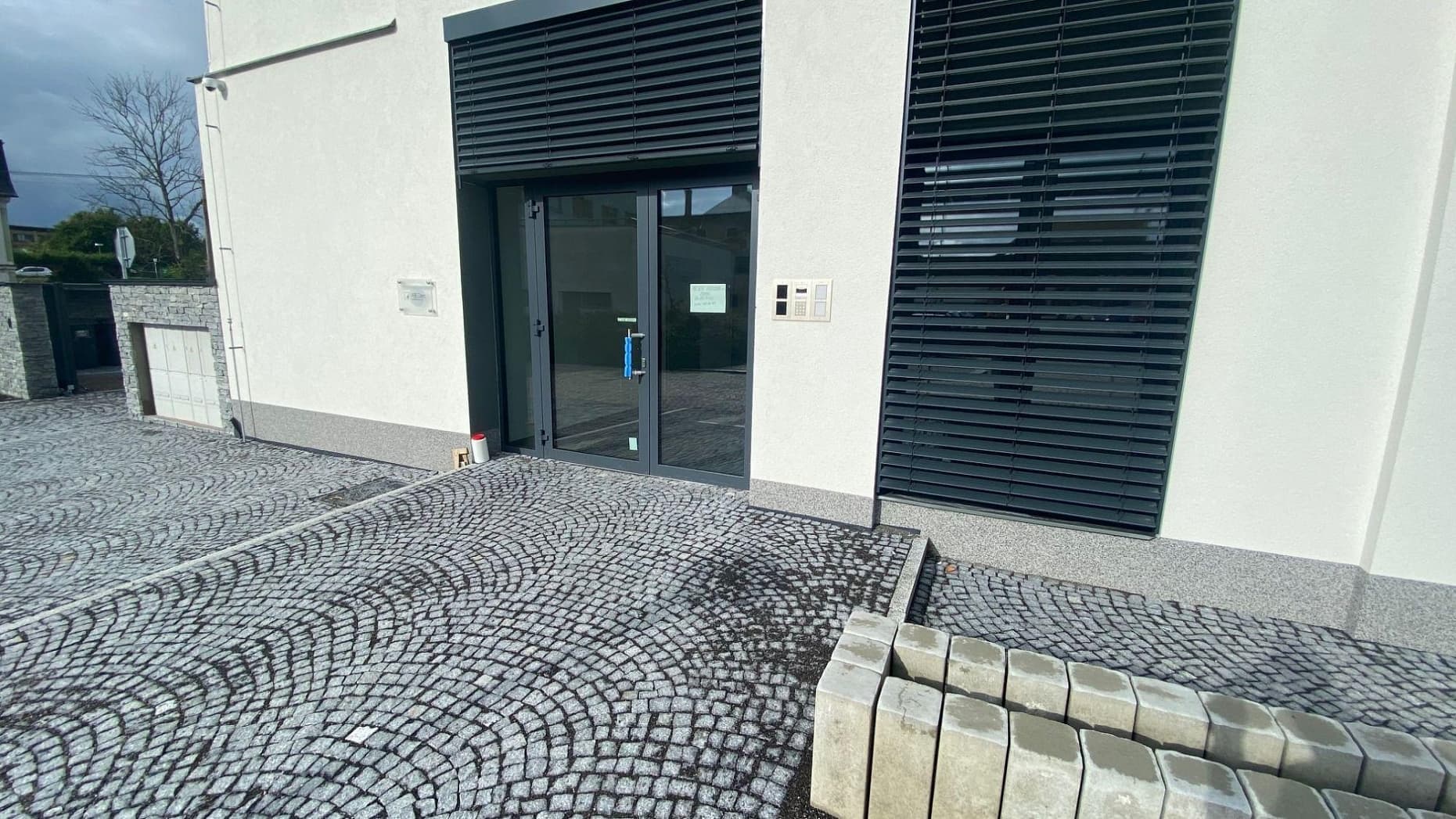 Vstup do moderní budovy s tmavou žulovou dlažbou v obloukovém vzoru, doplněnou kamenným obkladem.