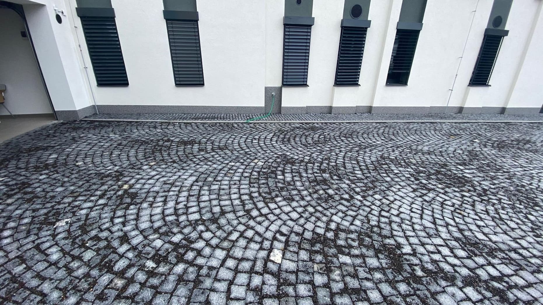 Dlážděná plocha z tmavé žulové kostky v obloukovém vzoru u moderní budovy s bílou fasádou a tmavými okenicemi.