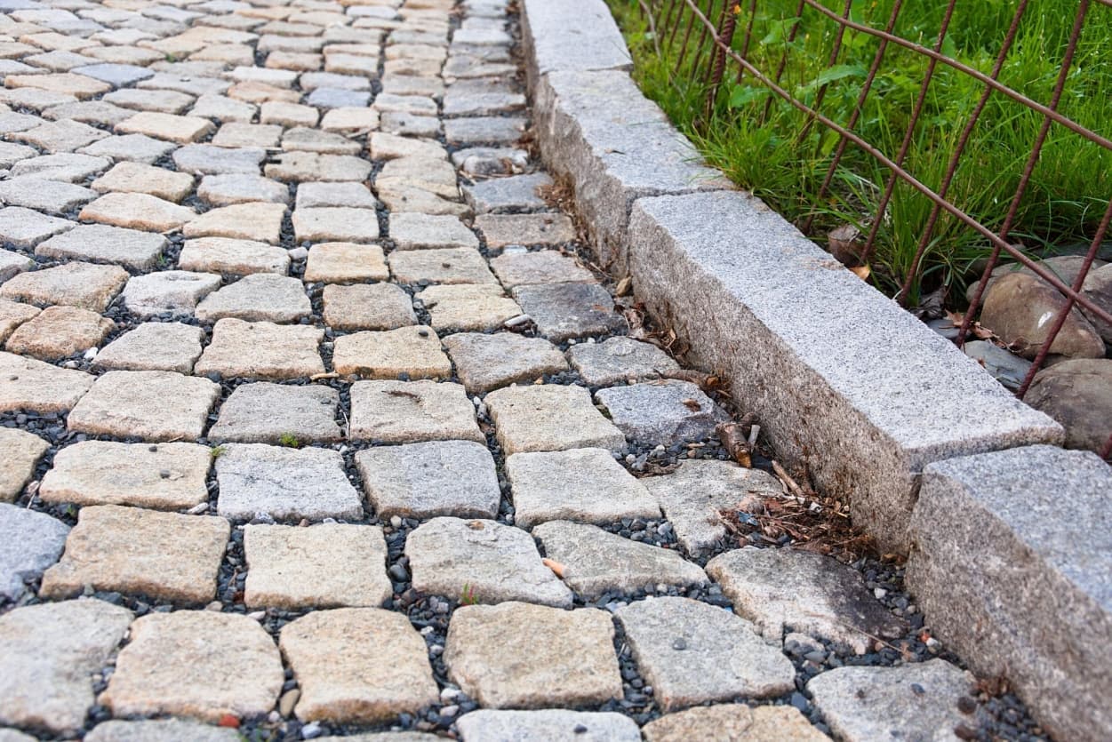 Detail žulových štípaných kostek ve žlutošedém odstínu s pevným žulovým krajníkem a štěrkovou výplní mezi dlažbou.